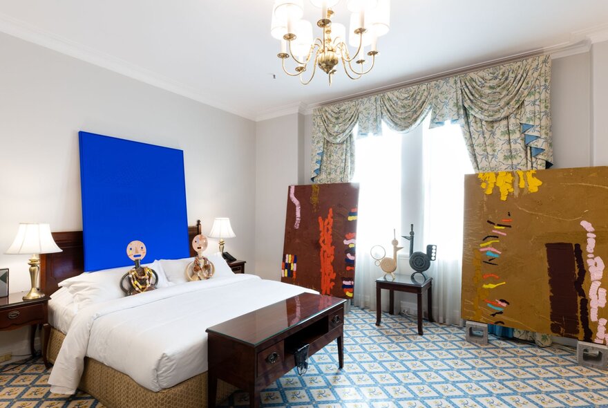 A room at the Hotel Windsor decorated with art works hanging on the walls or resting on the floor, and with two sculptural figures lying on the hotel bed.