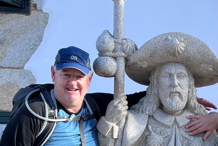A middle-aged man wearing hiking gear posing with a statue outdoors.