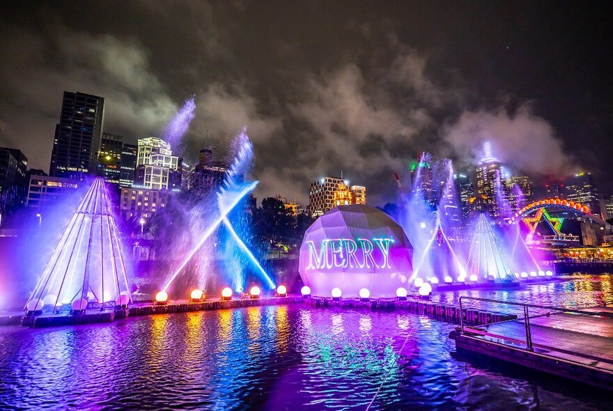 A light show on a river at night with water spouts and a bauble that reads 'merry'.