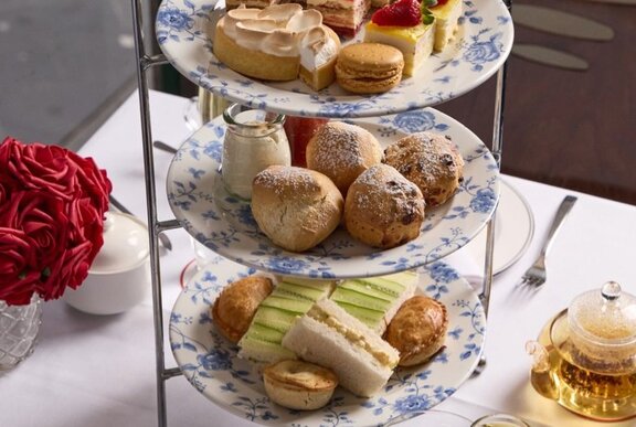 A high tea three-tiered plate of ribbon sandwiches, scones and sweets on a table, with a pot of tea and a vase of flowers.