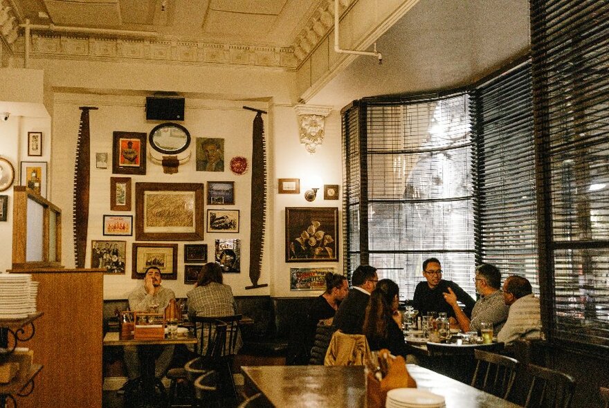 Interior of venue with people seated at dark furniture, a large window and rear wall displaying artwork of assorted sizes.