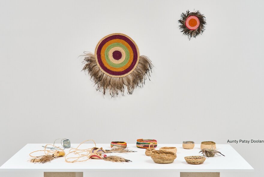 Textiles and woven objects hanging on a white wall, with woven objects on a low table display in the white gallery space at Craft Victoria.