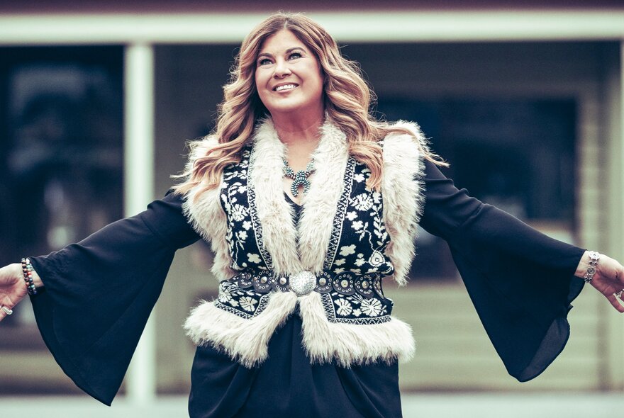 Singer/songwriter, Sandy Facee, standing with arms outstretched, wearing a black top with flared arms and a black and white sherpa vest.