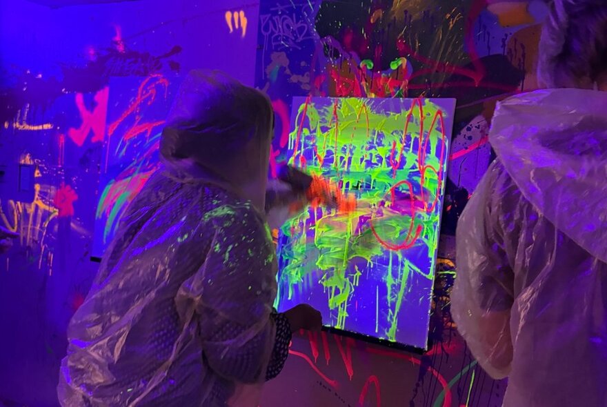 Two people in protective plastic clothing painting onto canvases with neon paints that are lit up with ultraviolet lights. 