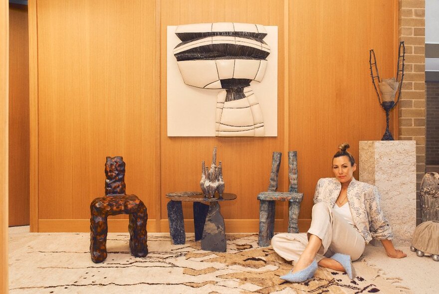 A stylish woman sitting on the floor next to three rugged looking ceramic sculptures in front of a timber-lined wall hung with a feature artwork.