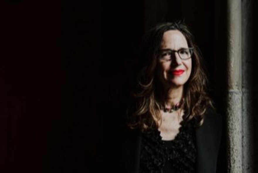 Author Elise Valmorbida, a middle-aged woman with long dark hair, wearing glasses, leaning against a wall in a shadowy recess.
