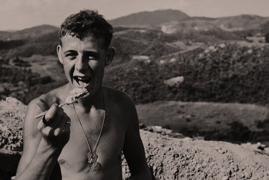 Black and white photograph of a shirtless soldier eating something from a spoon, trenches and hills seen behind him.