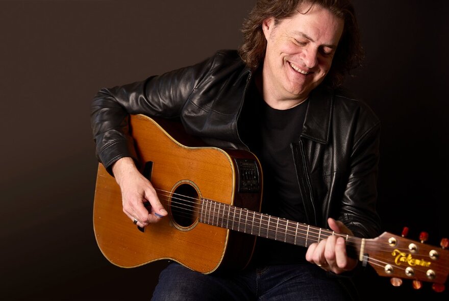 Musician smiling and playing an acoustic guitar, wearing a leather jacket.