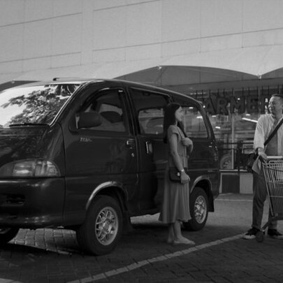Black and white film still, a couple with a trolley next to a car in a supermarket car park.