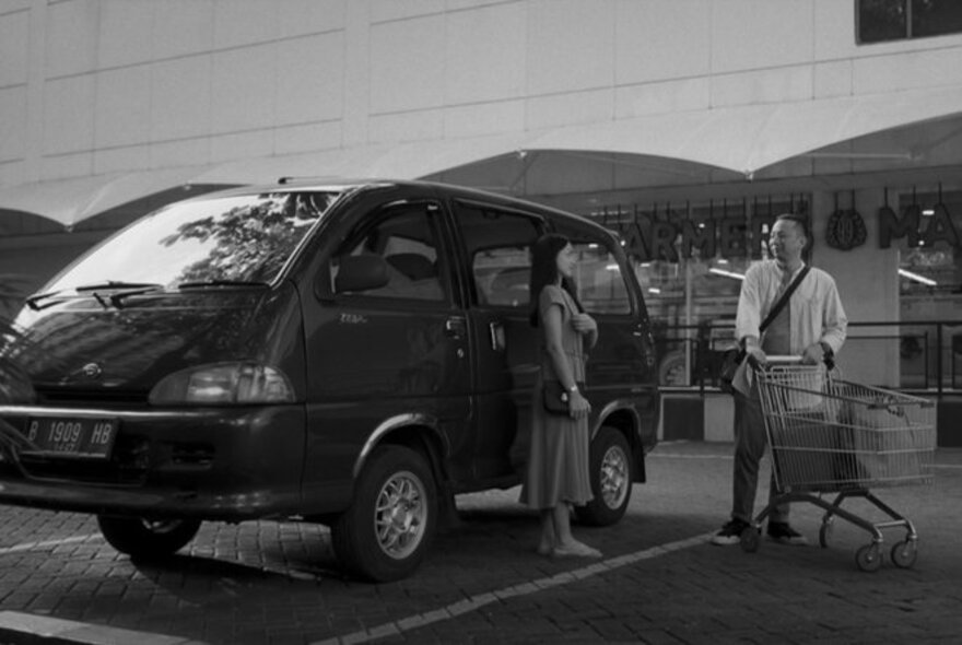 Black and white film still, a couple with a trolley next to a car in a supermarket car park.