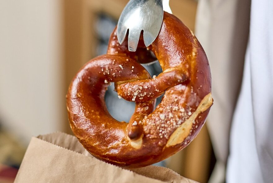 A pretzel pastry being placed into a paper bag with tongs.