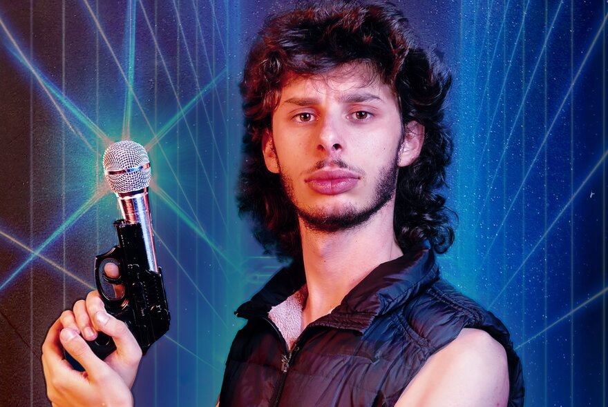 Comedian Con Coutis, a young man with curly hair and a beard, holding a microphone in his hand that has a base resembling a gun, posing in front of a blue background with receding grid lines on it.