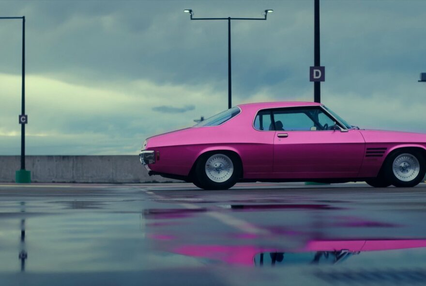 A hot pink muscle car on a rooftop carpark, the car reflected in a puddle of water. 