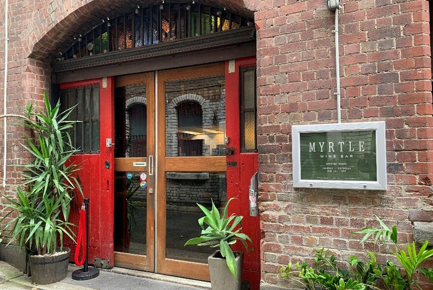 The brick exterior of a bar with red doors and plants outside.