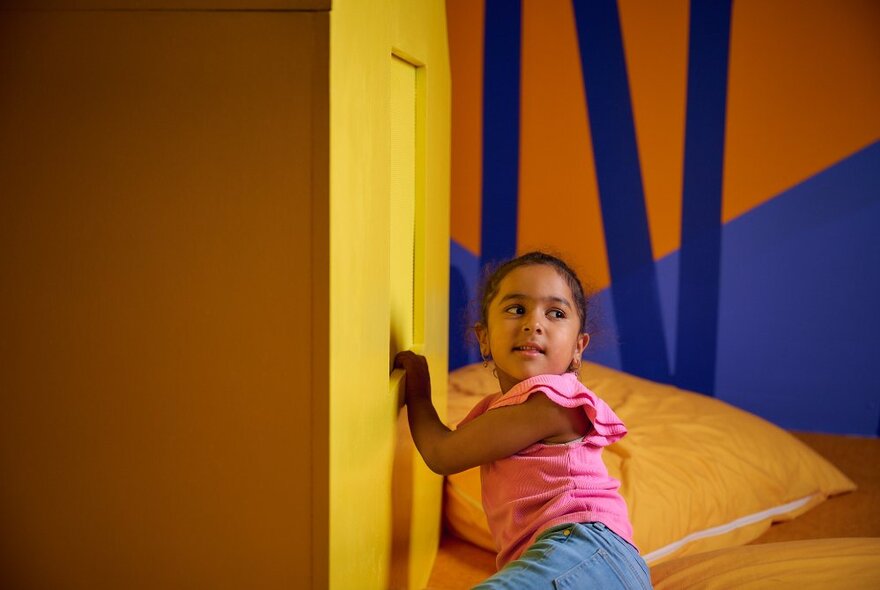 A young child in a brightly-coloured play space with cushions and a small door in the wall.