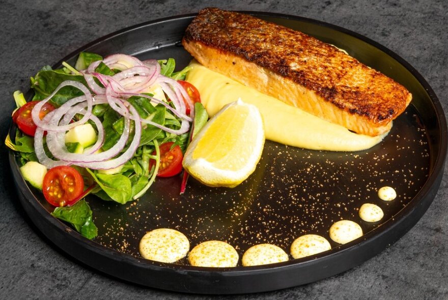 A slice of grilled fish on a black plate with circles of mayonnaise and salad.