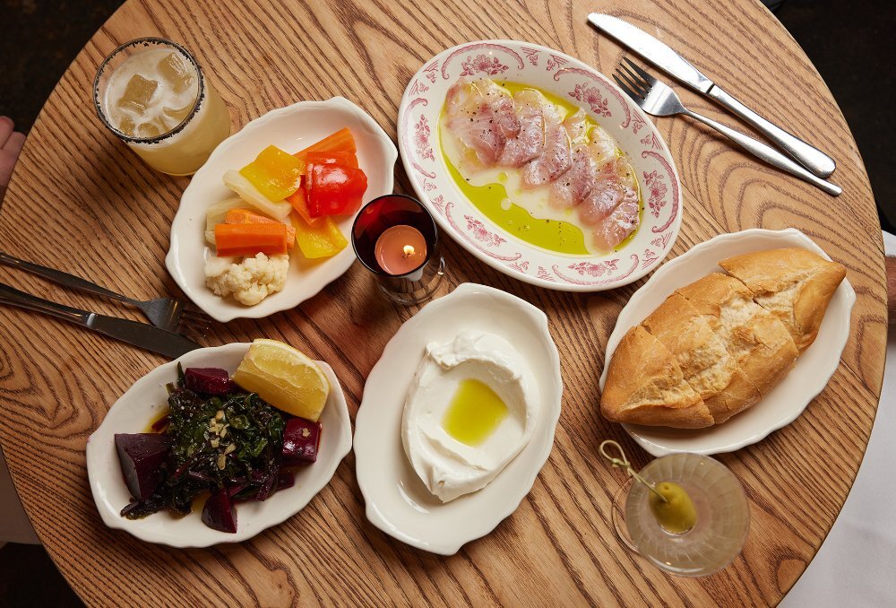 A table of Greek dishes including bread, dips and pickles.