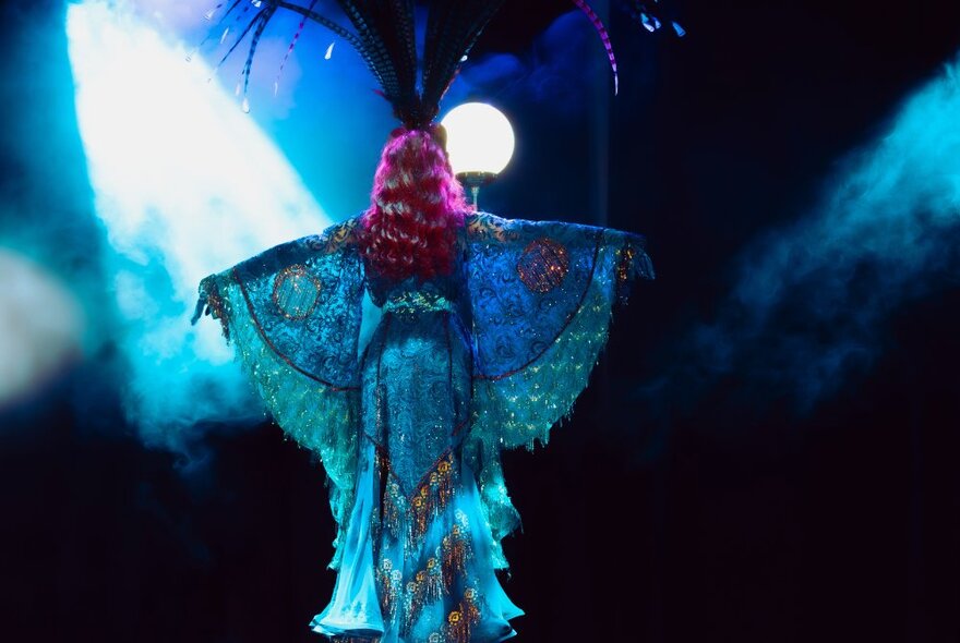 The back of a burlesque dancer with a tall headdress and long red hair performing with spotlights behind her. 