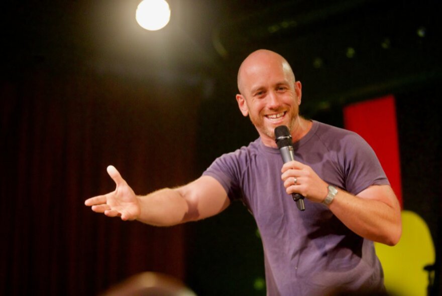 Danny McGinlay performing stand up comedy on a stage, holding a microphone in his hand.