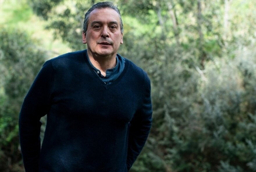 Author Christos Tsiolkas standing outside in a garden, wearing a navy jumper, with short greying hair.