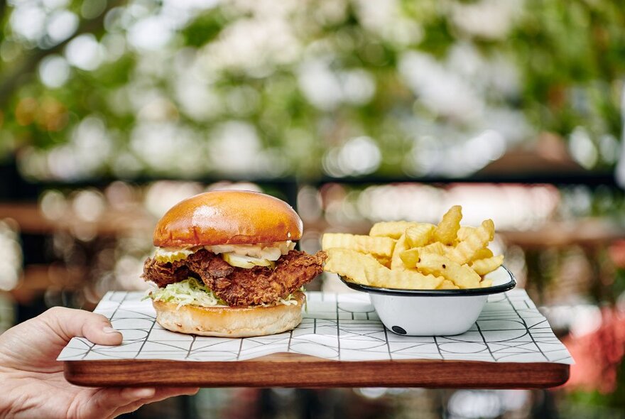 A chicken burger and crinkle cut chips on a wooden tray at an outdoor bar.