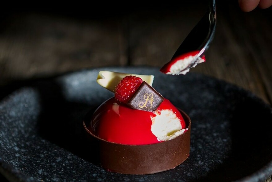 Close up of a small cheesecake with chocolate around bottom, covered in red with a raspberry and chocolate shards on top.