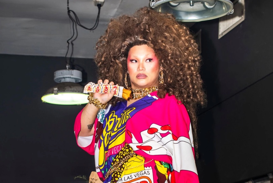 Portrait of drag queen, Aysha Buffet, with big hair, full makeup and a pink patterned robe-style dress, holding a Chiko Roll.
