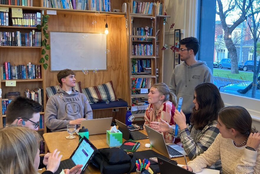 Teenagers sitting and working around a large table in a room, some with laptops open in front of them, bookshelves in the background and windows looking out onto the street on a side wall.