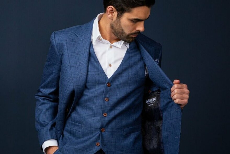 Man in blue suit, and white shirt, looking into his left inner coat pocket, against dark blue mottled background.