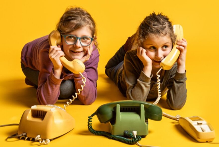 Two primary school-aged children kneeling on a yellow floor, interacting with vintage rotary telephones.
