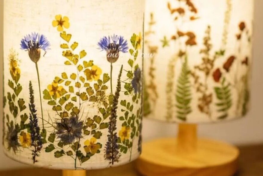 Two illuminated lamps with cylindrical parchment or fabric shades, decorated with various pressed flowers.