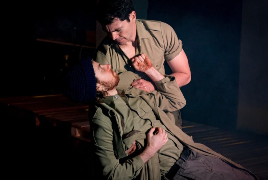 Two actors on stage, both dressed in soldier's clothes, one wounded and lying in the arms of the other who is comforting him.