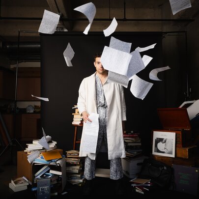 A man in a white lab coat stands in a dimly lit, cluttered studio as sheets of white paper fly and float through the air all around him.
