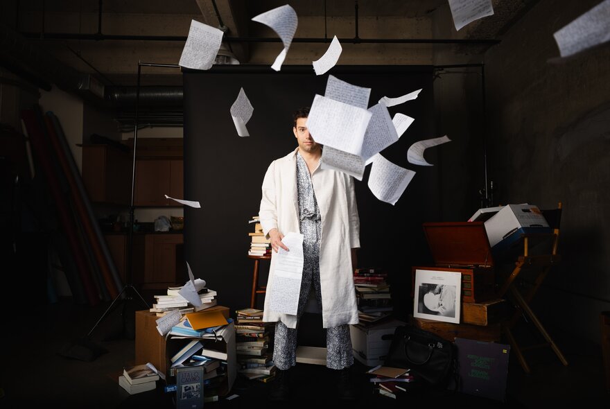A man in a white lab coat stands in a dimly lit, cluttered studio as sheets of white paper fly and float through the air all around him.