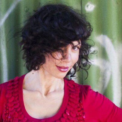 Singer Jackie Bornstein, one eye hidden by her dark hair, wearing a red frilled top, standing in front of green graffitied corrugated iron fence.
