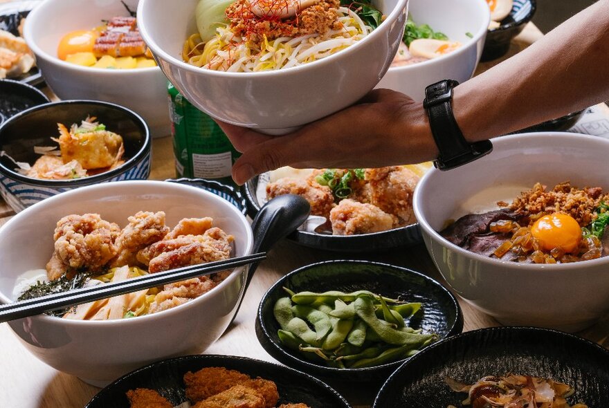 Japanese noodle dishes displayed on a wooden table, with a hand picking up a bowl of ramen.