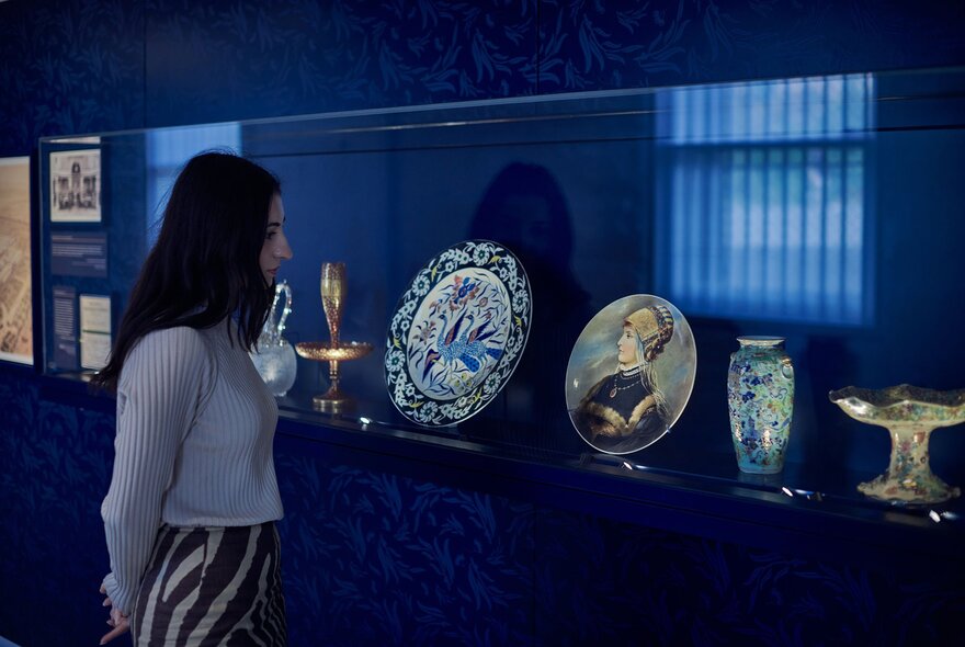 A person with long dark hair and their hands behind their back looking at valuable ceramic images inside a glass display case, set against a dark blue wall.