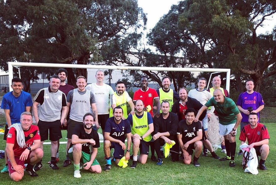 A group of mostly men of varied ages, posing in their athletic wear, in front of a soccer goalie net at an outdoor oval.