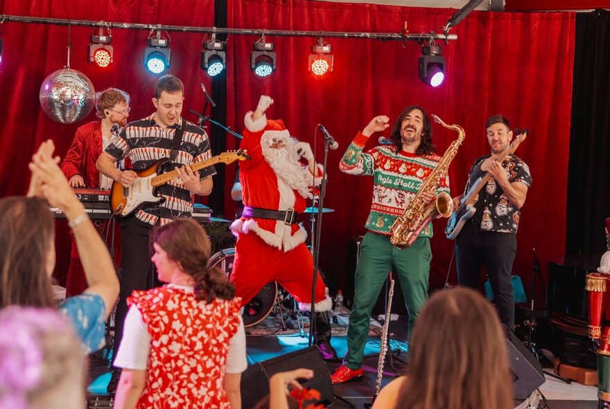 A festive holiday music performance featuring a band playing live on stage decorated with red curtains, disco balls, and a person in a Santa Claus suit, with some children standing at the front watching the show.