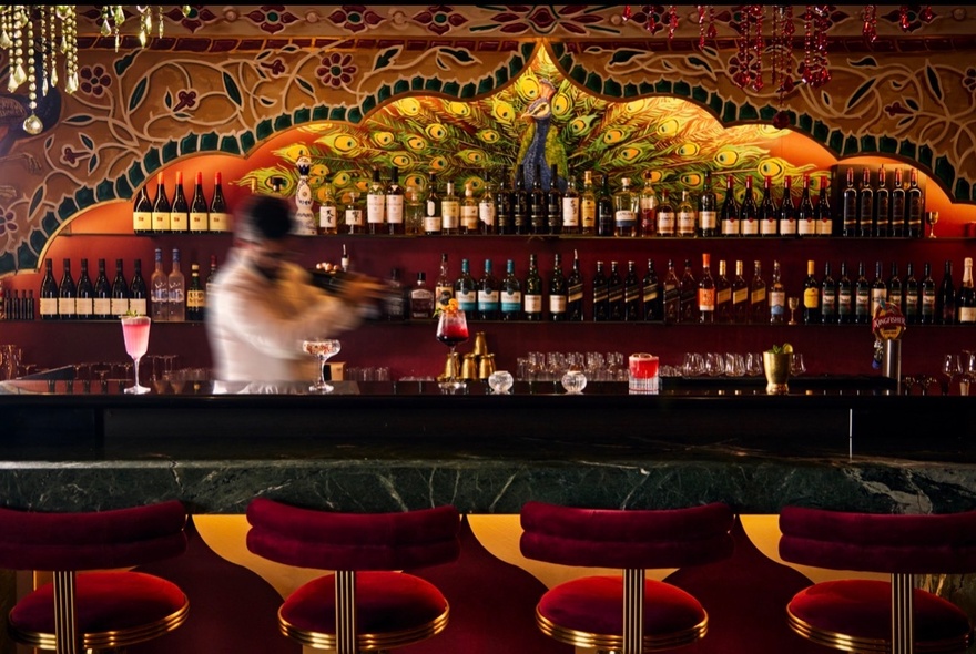 Indian-themed bar with peacock design, blurred barman blending cocktails, red velvet barstools.