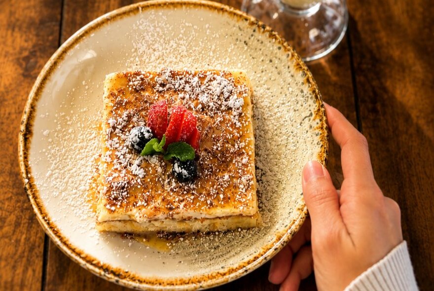 Top-down view of a thick slice of dessert on a rustic ceramic plate, dusted with powdered sugar and topped with fresh berries and a sprig of mint.