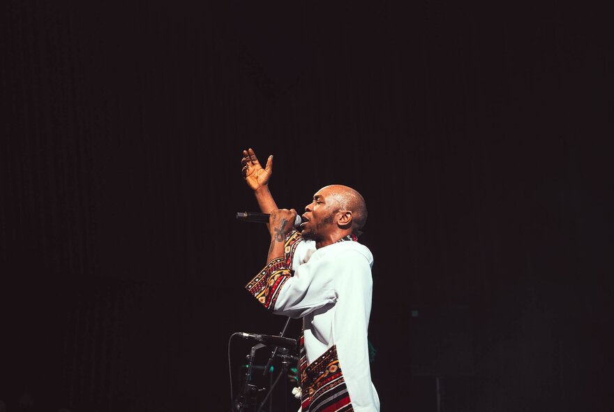 Musician Sean Kuti in profile, singing into a microphone on a darkened stage, one arm raised in the air above his head.