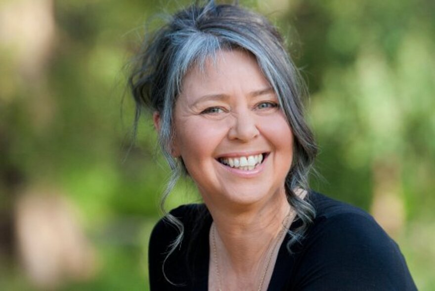 A portrait of a smiling woman with tied-up greying hair, with blurred greenery in the background.