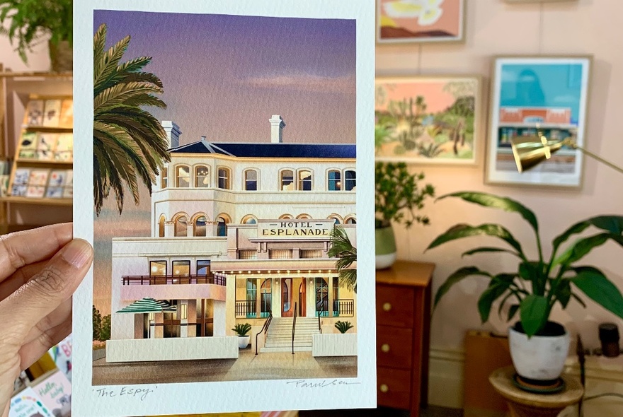 A hand holding up a drawing on paper of St Kilda's Hotel Esplanade, behind this drawing is the interior of a gift shop with shelves against the wall, plants and pictures hanging on the wall. 