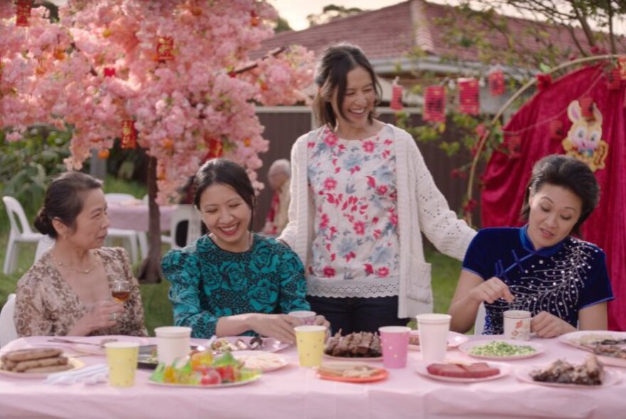 A long table set with a pink tablecloth and snacks and drinks, with four women  and a cherry blossom tree behind. 