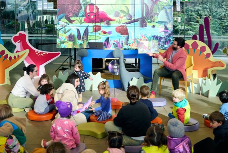 Young children and their parents listening to a story book being read aloud in a colourful environment with large dinosaur cutouts.