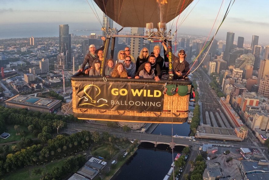 A group of people in a hot air balloon basket floating above Melbourne, Go Wild ballooning signage.