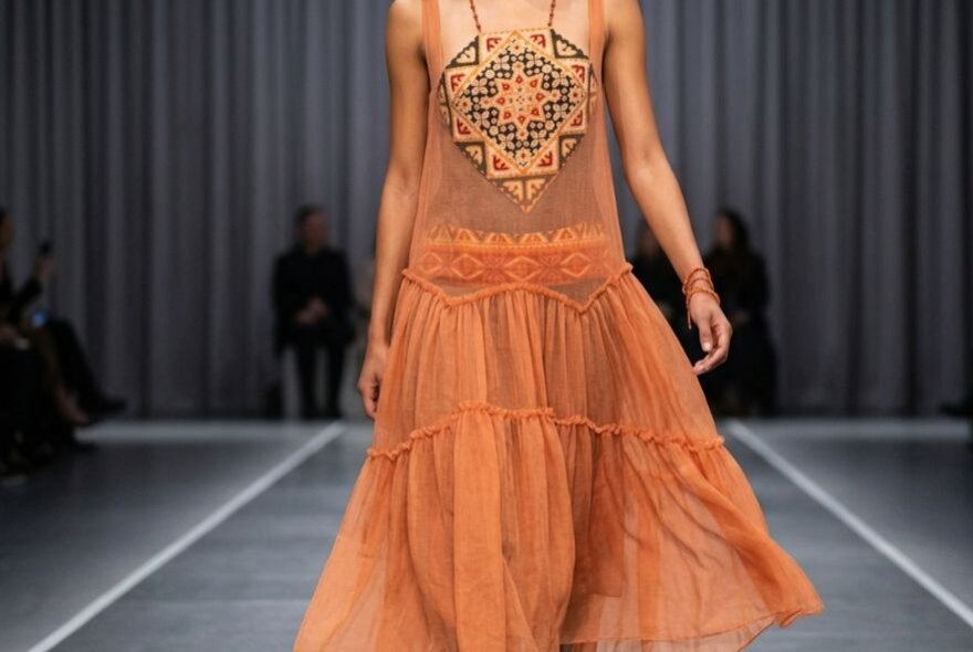 A model wearing a burnt-orange sheer tulle maxi dress on a runway, made from recycled and upcycled fabric.