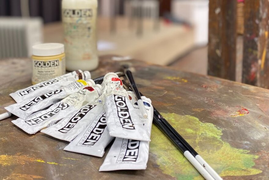 Tubes of acrylic paint on a table with two paint brushes.