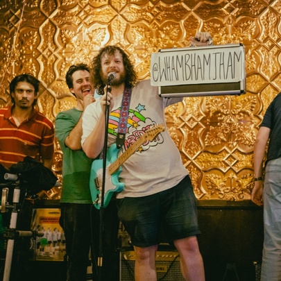 Four people on a stage, two of them with instruments, and one of them holding up a hand written sign that reads WHAMBHAMJHAM.